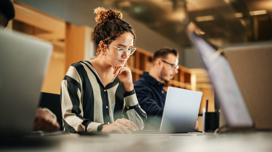 Portrait Of Enthusiastic Hispanic Young Woman Working On Computer In A Modern Bright Office. Confident Human Resources Agent Smiling Happily While Collaborating Online With Colleagues.
